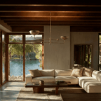 Clérachic brass chandelier glowing above a cream sectional sofa, framed by timber beams and a lakeside view.