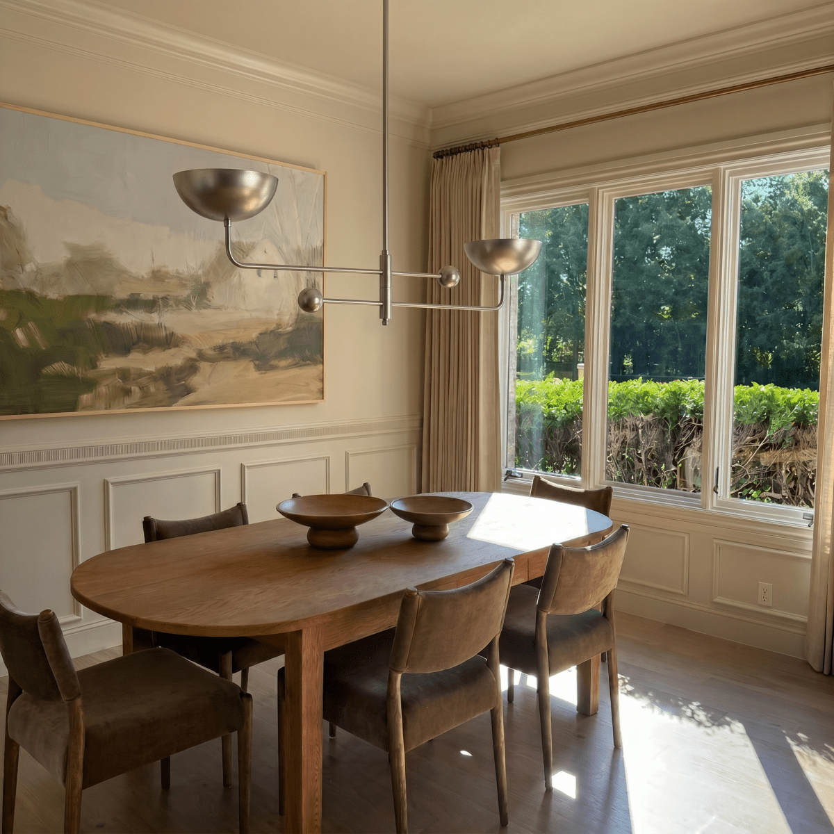 Clérachic chrome chandelier hanging above an oval dining table beside a landscape painting and garden windows.
