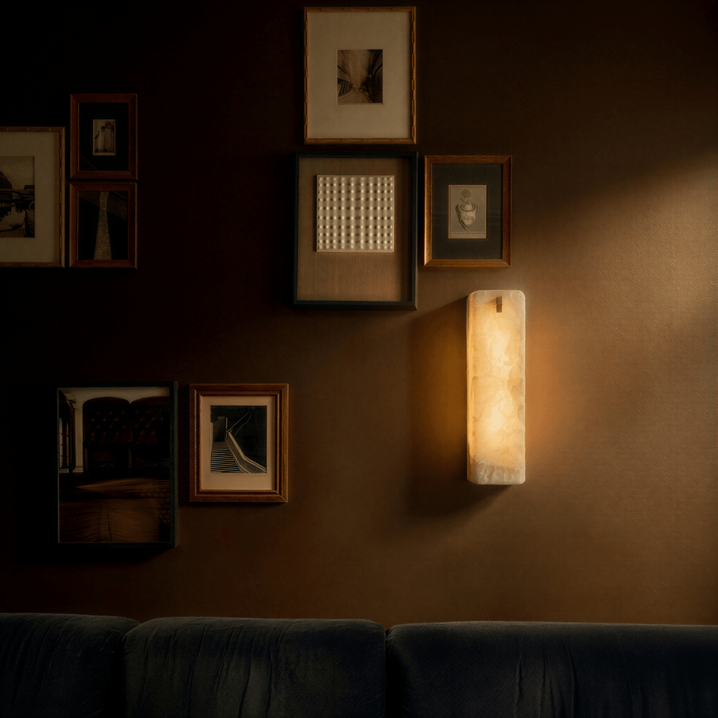 Clérachic marble wall sconce lit beside a gallery wall of small framed prints, its glow spilling onto the warm brown wall above a dark sofa.