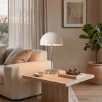 A chrome Clérachic table lamp lights a wood side table set with a book and a bowl of pistachios.