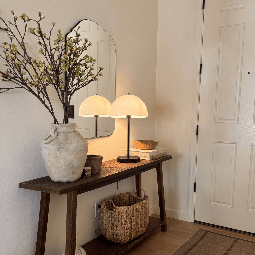 A black Clérachic table lamp lights a rustic console table beside a stoneware vase and woven basket.
