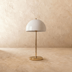 A polished brass Clérachic table lamp viewed from a different angle, the white dome shade lit warmly.