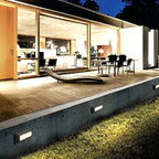 The lights set into a concrete retaining wall at night, washing the grass below a lit house.