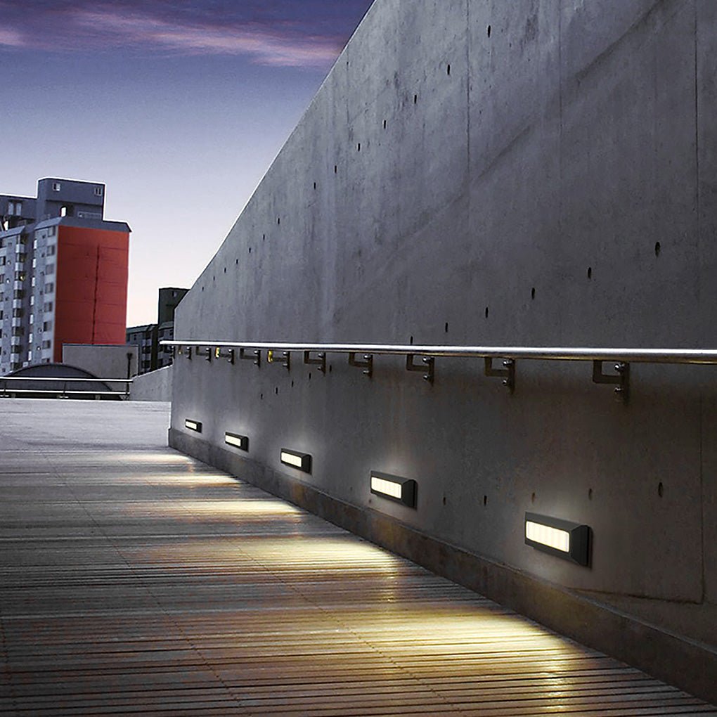 A row of the rectangular lights lit along a concrete walkway wall at dusk, city towers beyond.