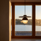 Clérachic pendant light in walnut glowing beside a window at dusk with a lake and mountains in view.