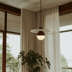 Clérachic pendant light in walnut glowing beside a tall window with a leafy potted plant nearby.