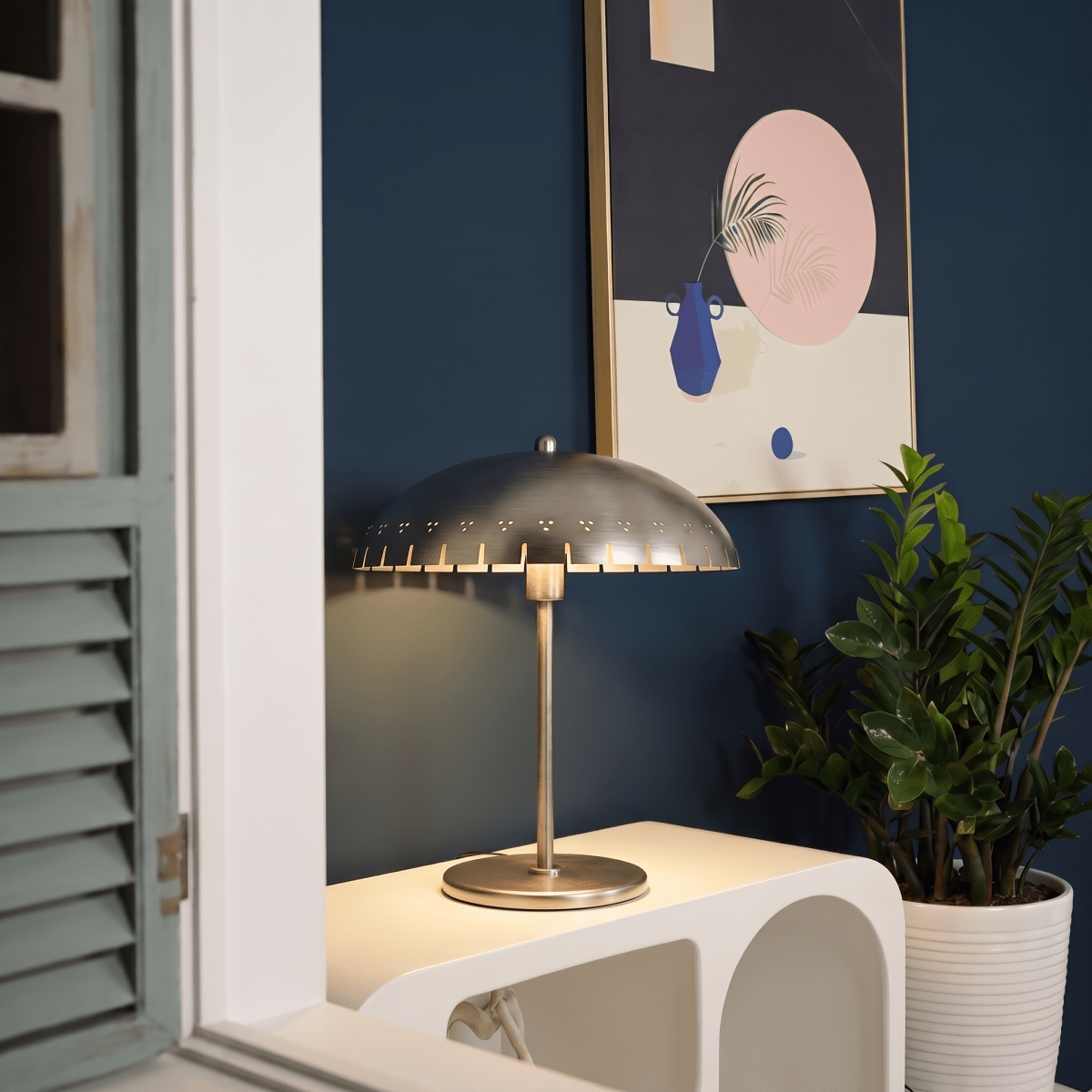 Clérachic table lamp in chrome glowing on a console beside shuttered doors and framed still-life art.