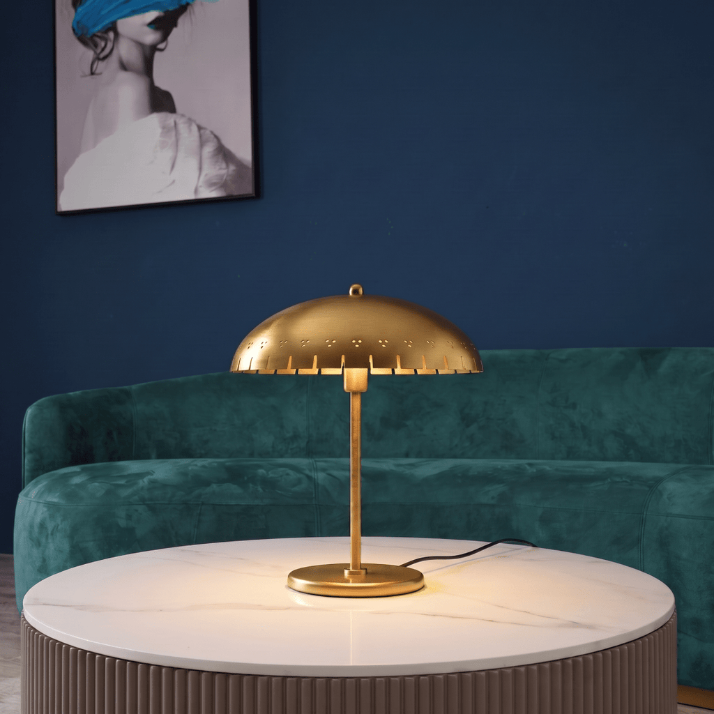 Clérachic table lamp in brass glowing on a marble table in front of a green velvet sofa and artwork.