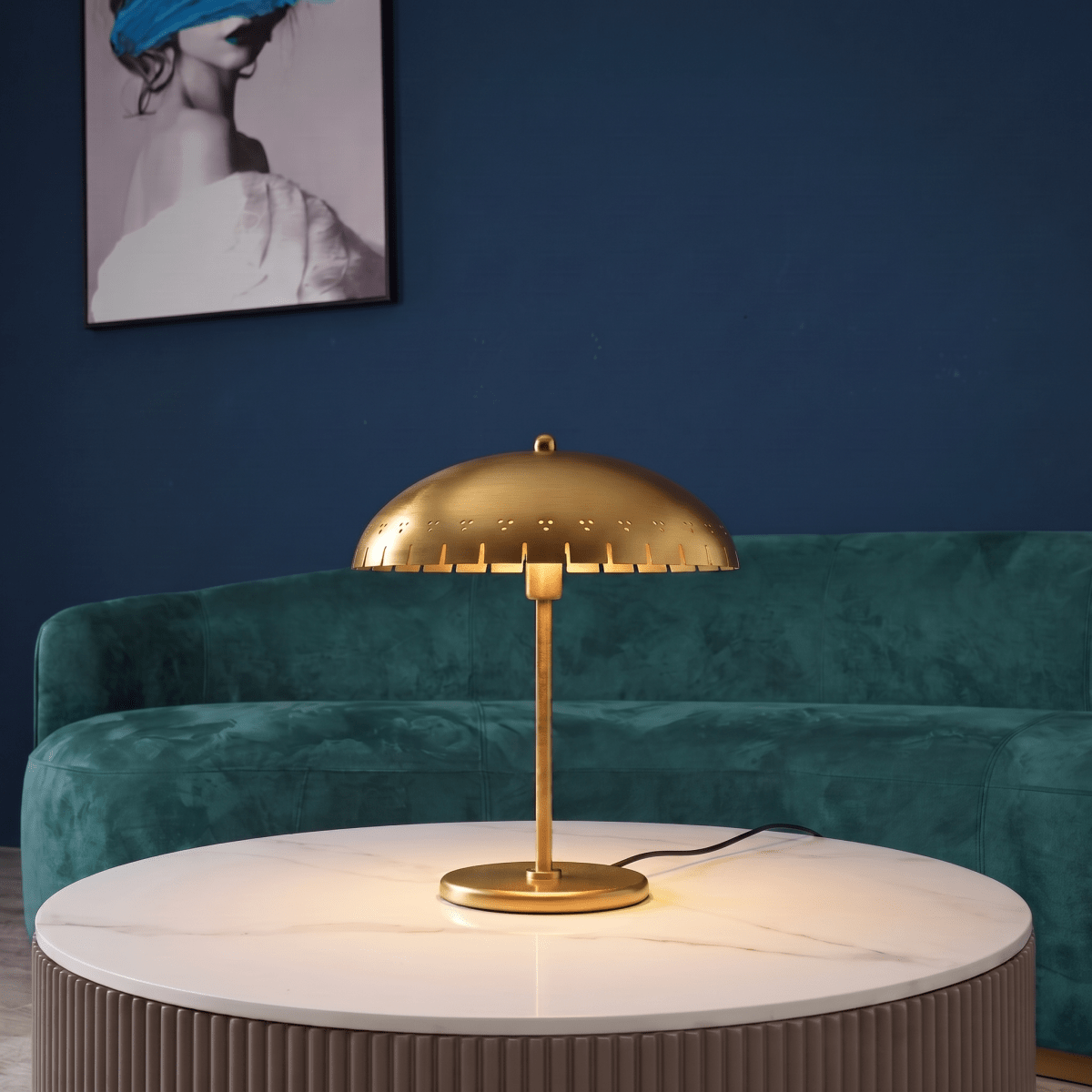 Clérachic table lamp in brass glowing on a marble table in front of a green velvet sofa and artwork.