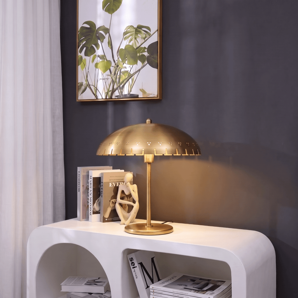 Clérachic table lamp in brass glowing on a console table beside stacked books and a monstera print.
