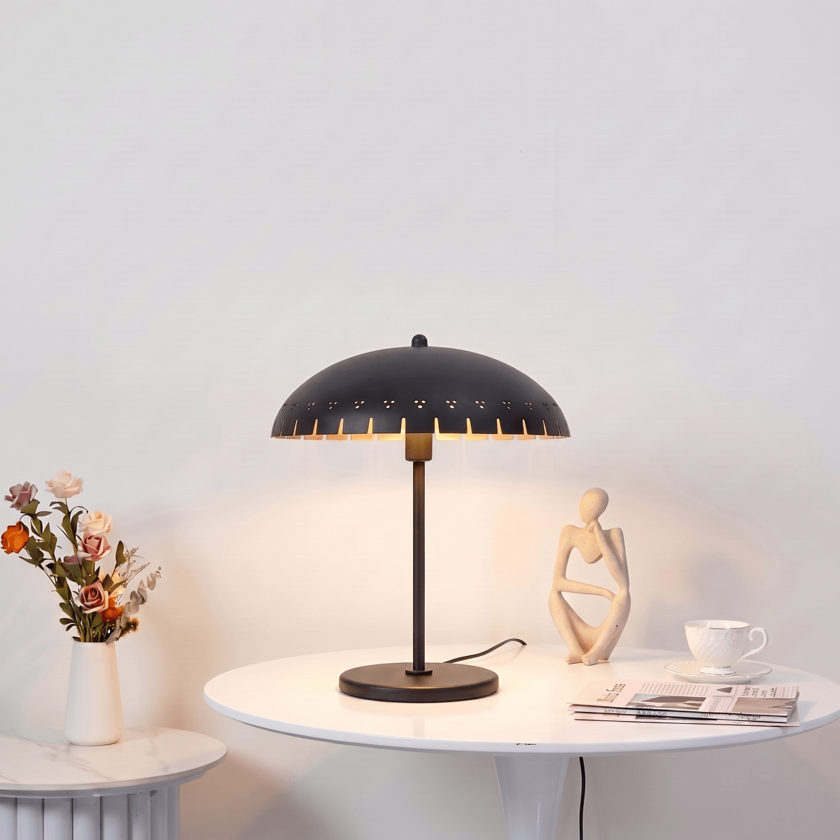 Clérachic table lamp in matte black glowing on a round white table beside roses and a small figurine.
