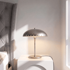 Clérachic table lamp in chrome glowing on a nightstand beside a tufted headboard and sunlit window.