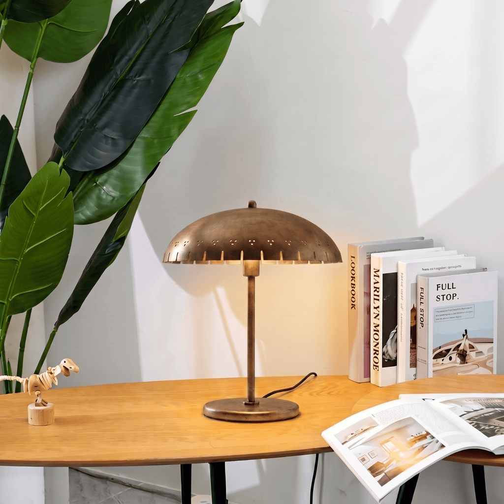 Clérachic table lamp in brass glowing on a wood table beside a stack of books and a toy dinosaur.