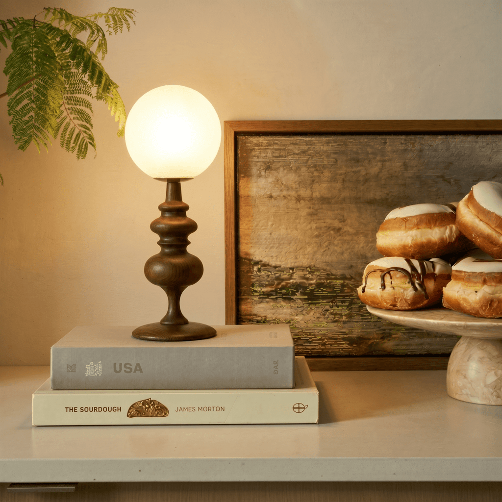 Clérachic table lamp in walnut glowing on a shelf beside a stack of books and a plate of pastries.