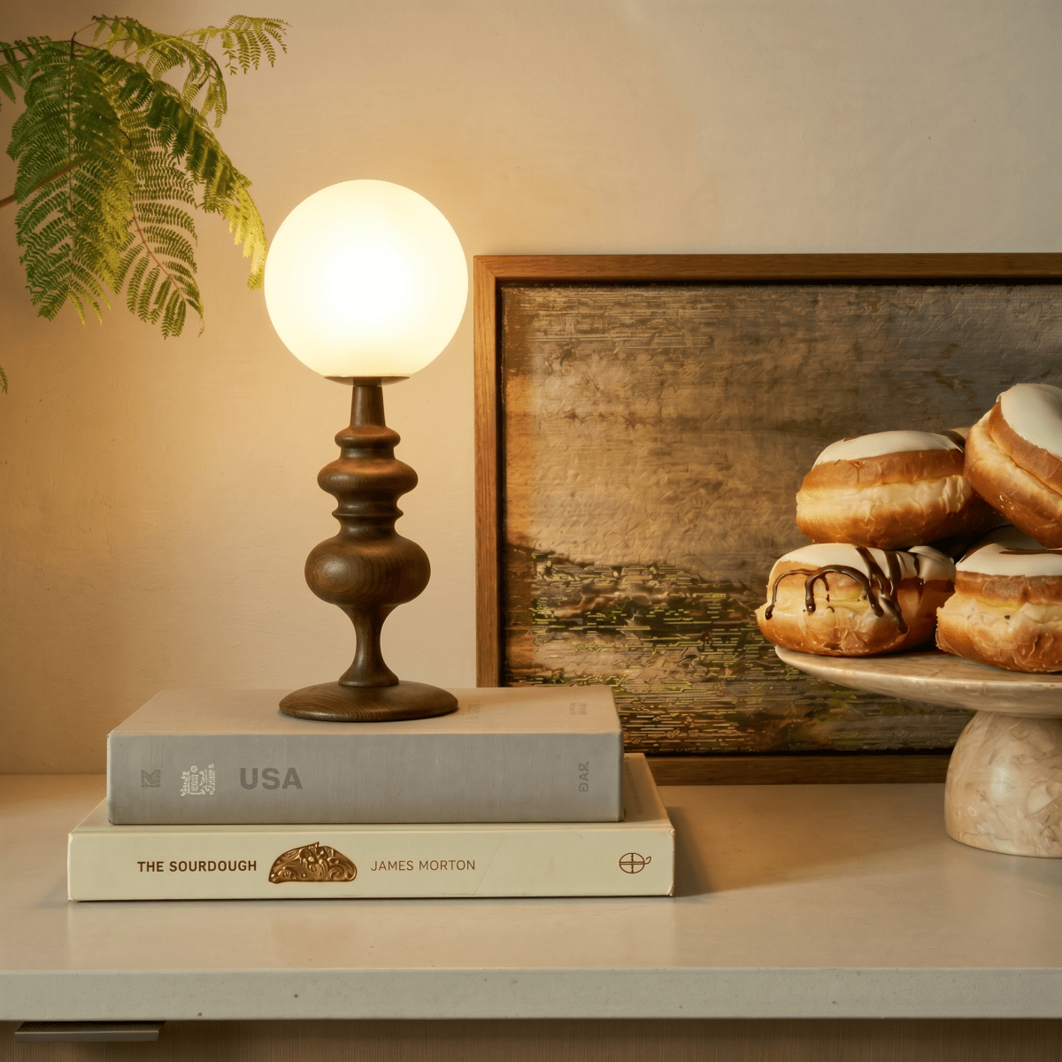 Clérachic table lamp in walnut glowing on a shelf beside a stack of books and a plate of pastries.