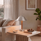 Clérachic table lamp in chrome on a travertine table beside a sofa, a book and a ceramic dish.