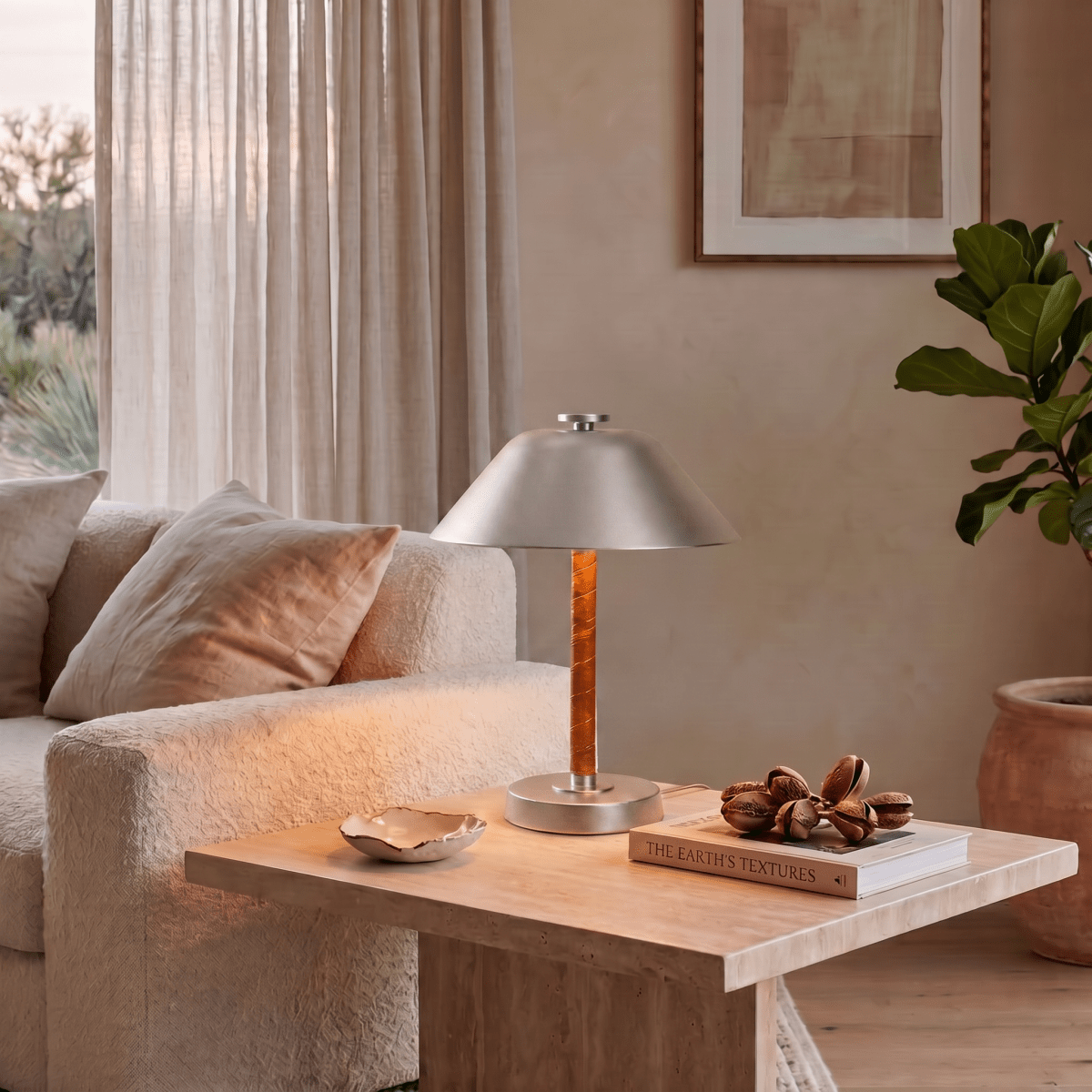 Clérachic table lamp in chrome on a travertine table beside a sofa, a book and a ceramic dish.