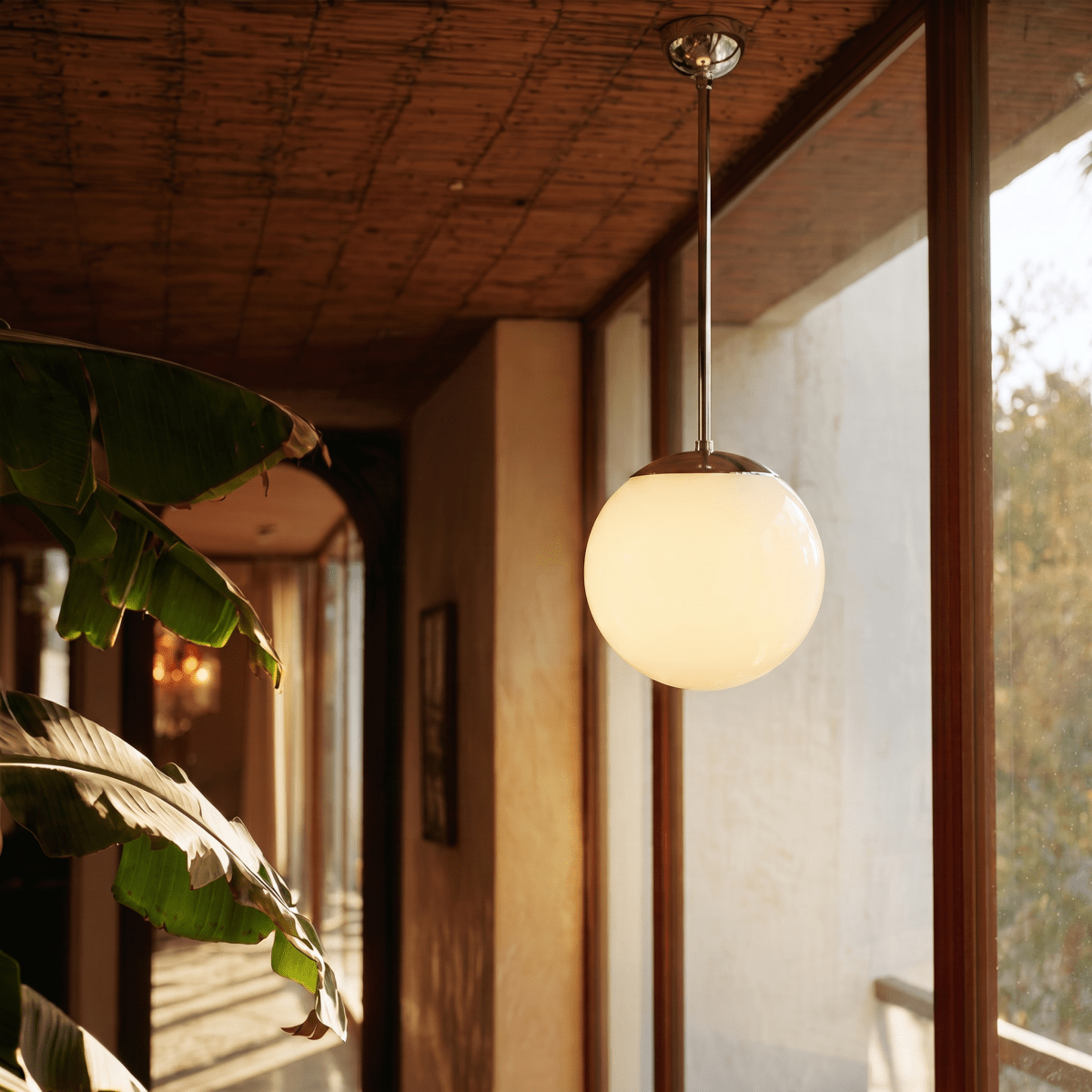Clérachic chrome globe pendant light lit on a covered terrace beside large tropical leaves at golden hour.