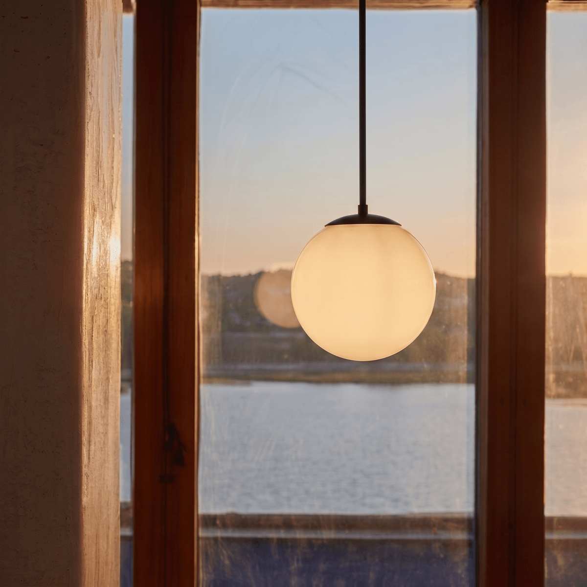 Clérachic matte black globe pendant light silhouetted against a window overlooking water at sunset.