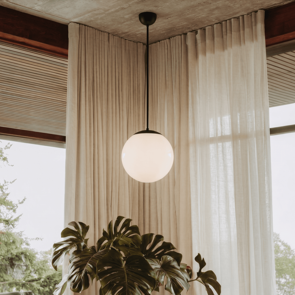 Clérachic matte black globe pendant light lit in a sunroom corner with sheer linen curtains and a monstera plant.