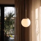 Clérachic brass globe pendant light lit beside a glass door with palm trees and warm evening light outside.