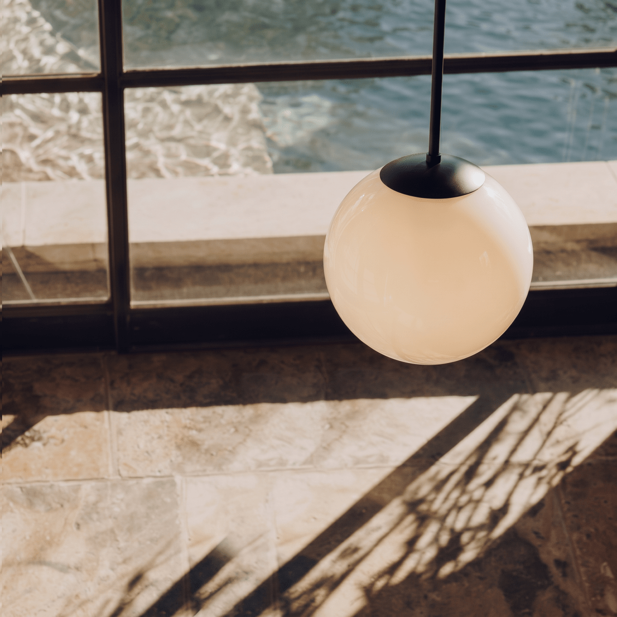 Clérachic matte black globe pendant light lit above a tiled terrace overlooking a pool in afternoon sun.