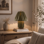 Clérachic table lamp in pleated olive green fabric glowing on a wood side table beside stacked books.