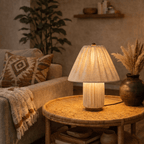 Clérachic table lamp in pleated beige fabric glowing on a rattan side table beside a patterned cushion.