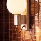 Clérachic wall lamp detail in travertine and brass, glowing against a dark tiled bathroom wall.