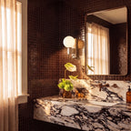 Clérachic wall lamp glowing beside a mirror in a dark tiled bathroom with a marble vanity below.