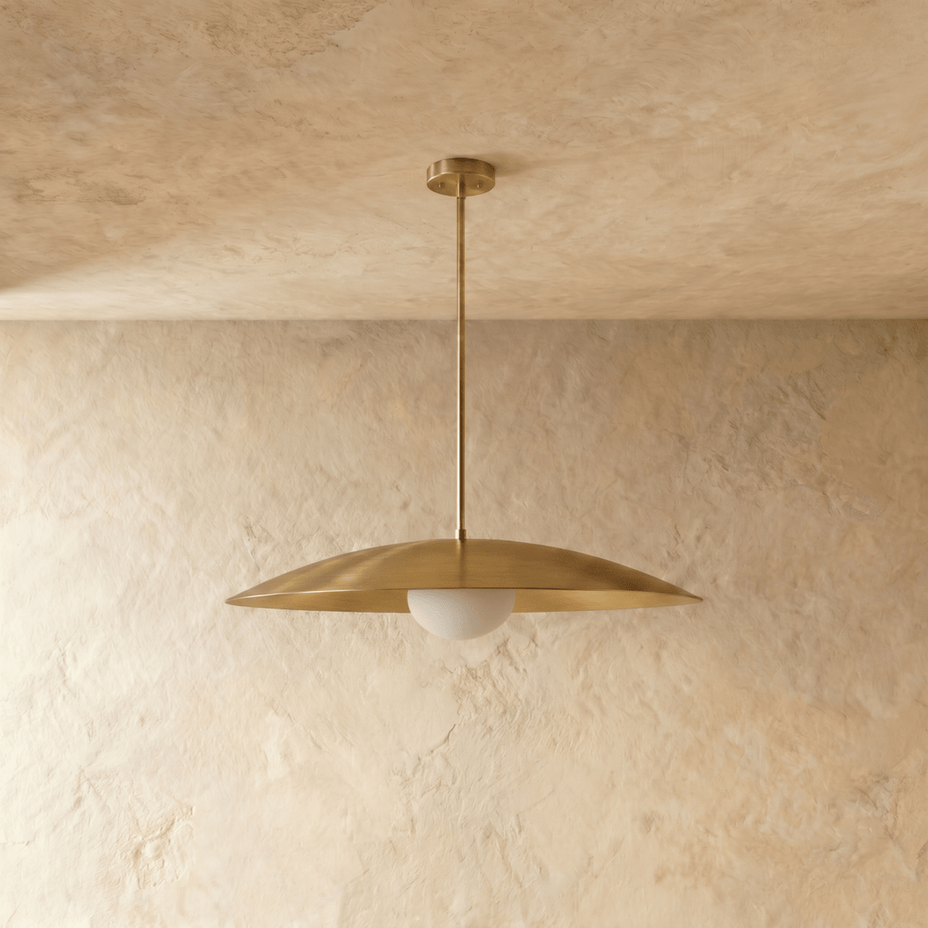 A polished brass Clérachic pendant light viewed from below, the opal diffuser glowing against a plaster wall.