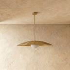 A polished brass Clérachic pendant light viewed from below, the opal diffuser glowing against a plaster wall.
