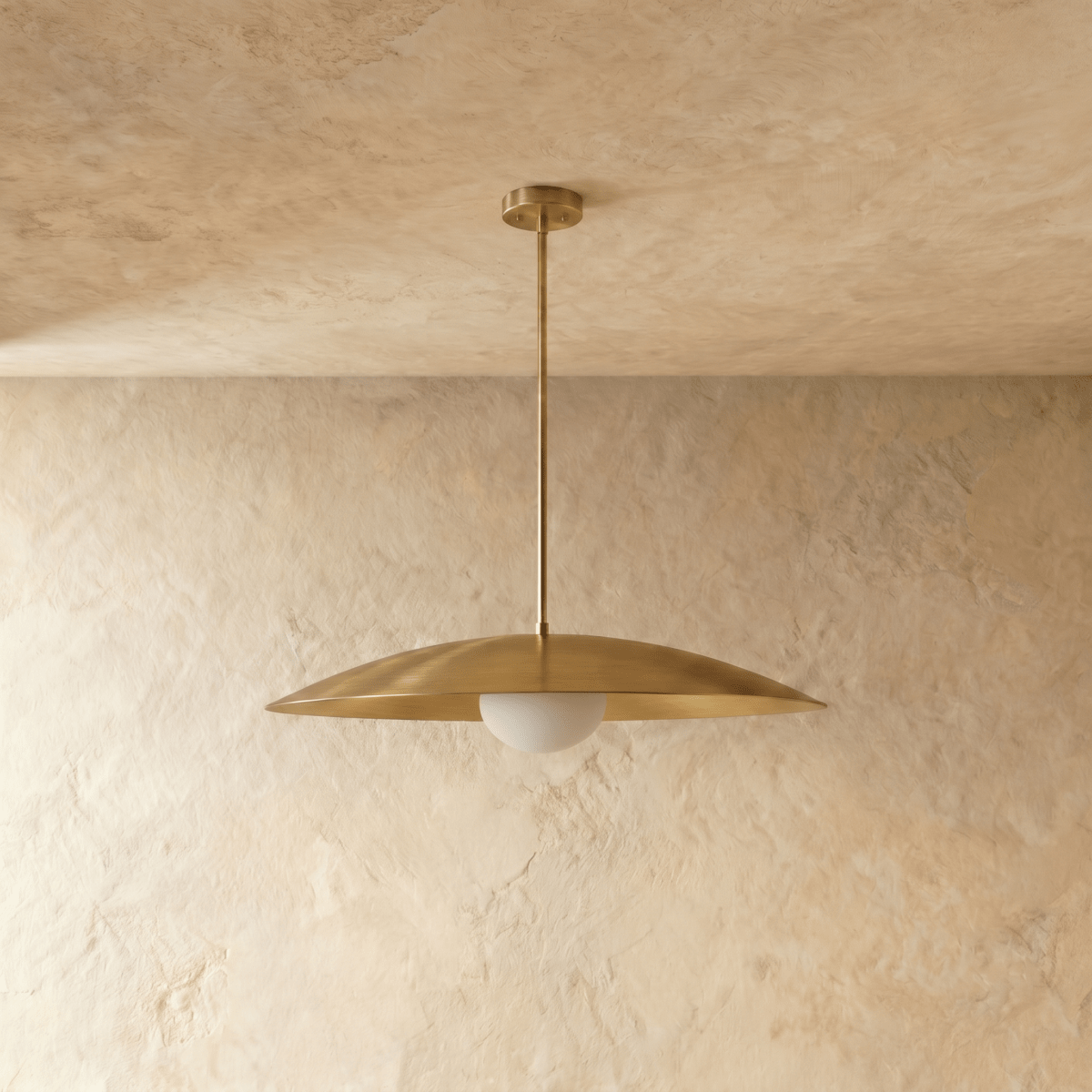 A polished brass Clérachic pendant light viewed from below, the opal diffuser glowing against a plaster wall.