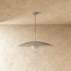 A chrome Clérachic pendant light viewed from below, the opal glass diffuser lit against a plaster wall.