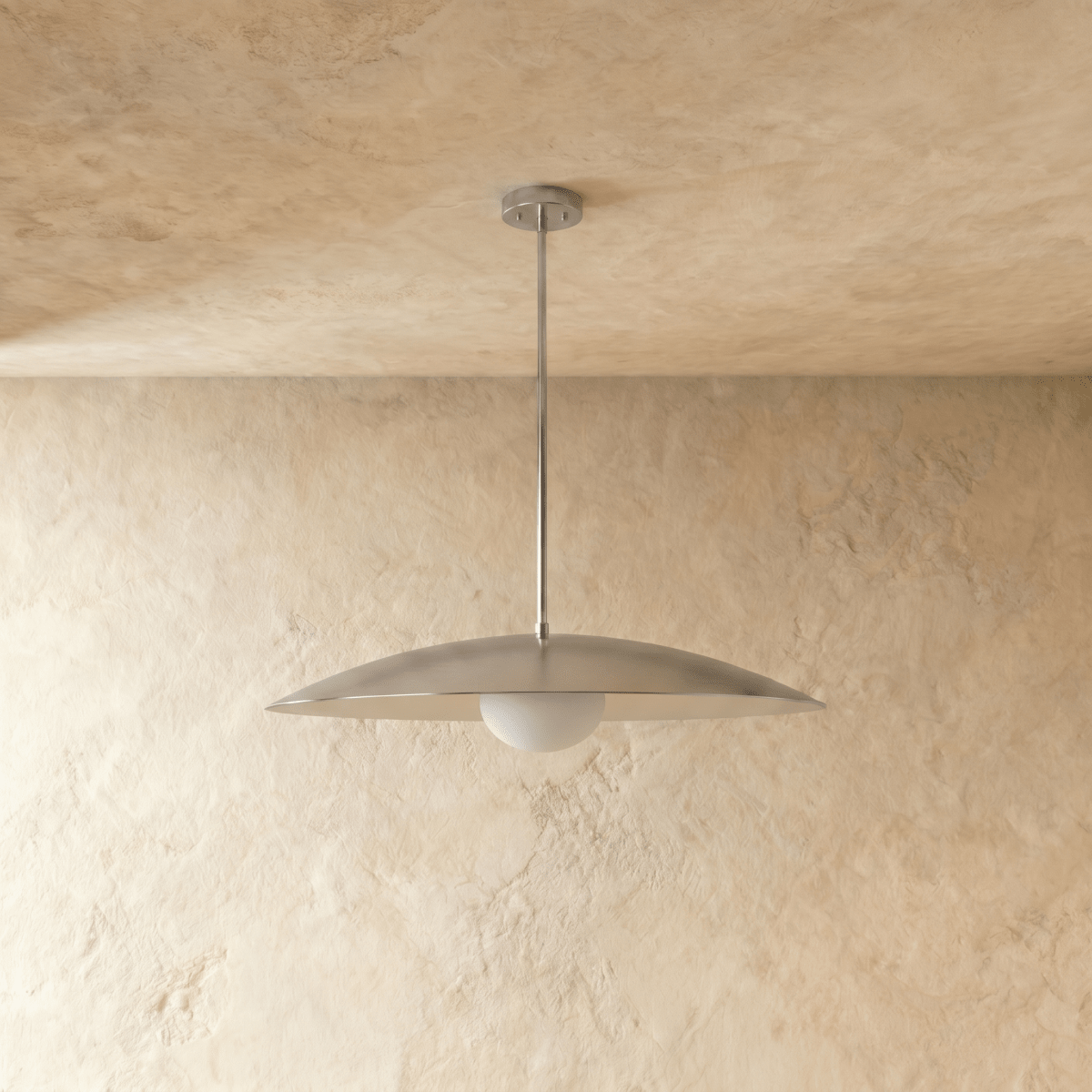 A chrome Clérachic pendant light viewed from below, the opal glass diffuser lit against a plaster wall.