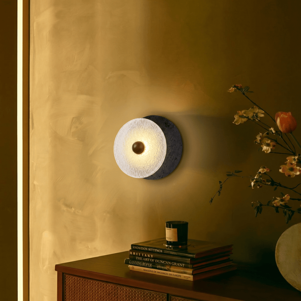 Clérachic wall lamp lighting a warm ochre wall above a cane-fronted sideboard stacked with books, a candle and a vase of flowering branches.