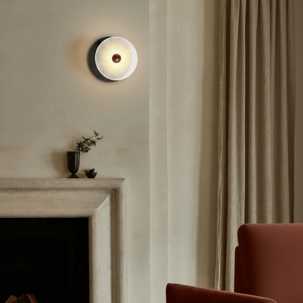 Clérachic wall lamp mounted high on a pale wall above a fireplace surround, its stone crescent showing to the left beside a floor-length curtain and a rust-coloured armchair.