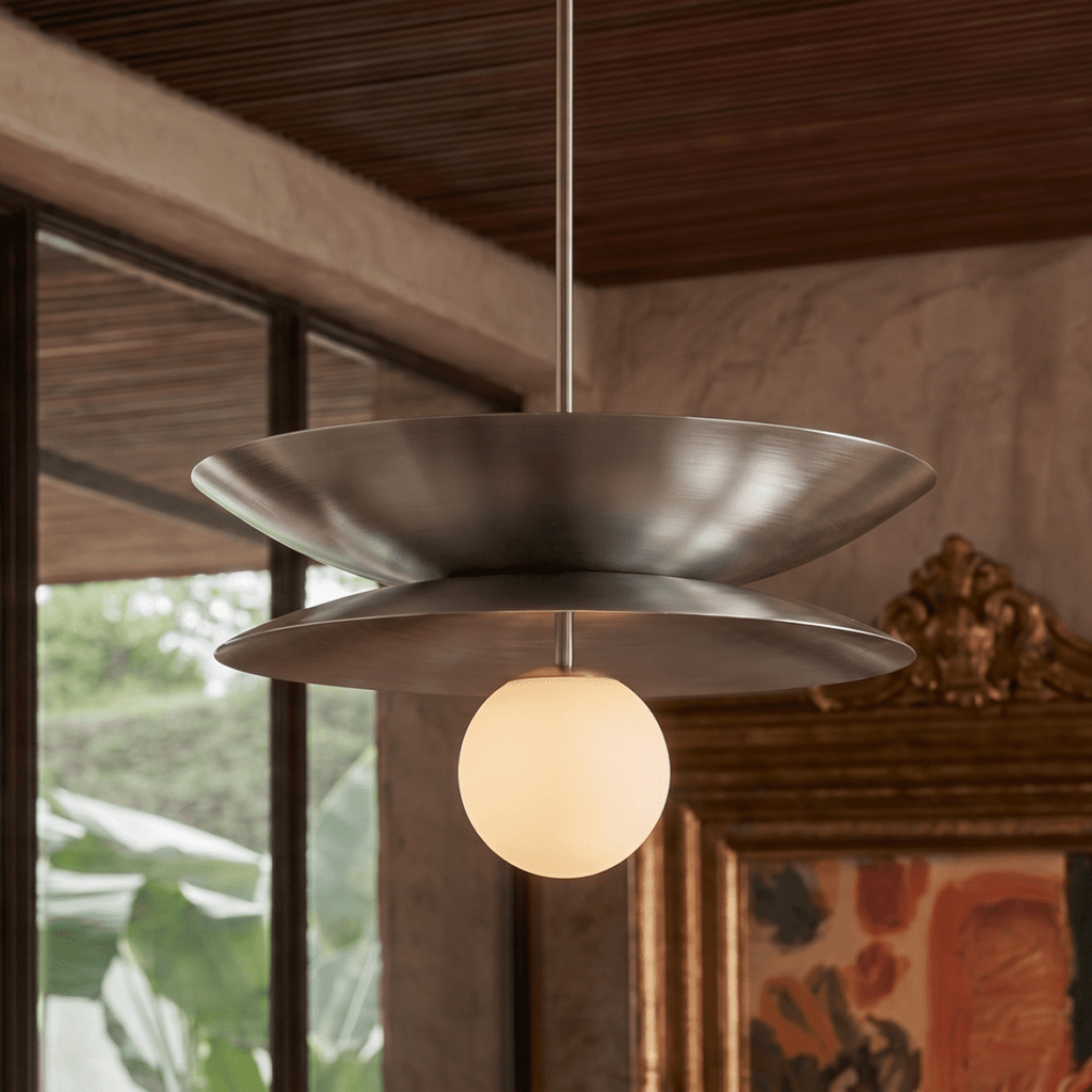 Clérachic layered-disc pendant light in chrome, its satin discs lit beside a gilt-framed painting with greenery beyond the window.