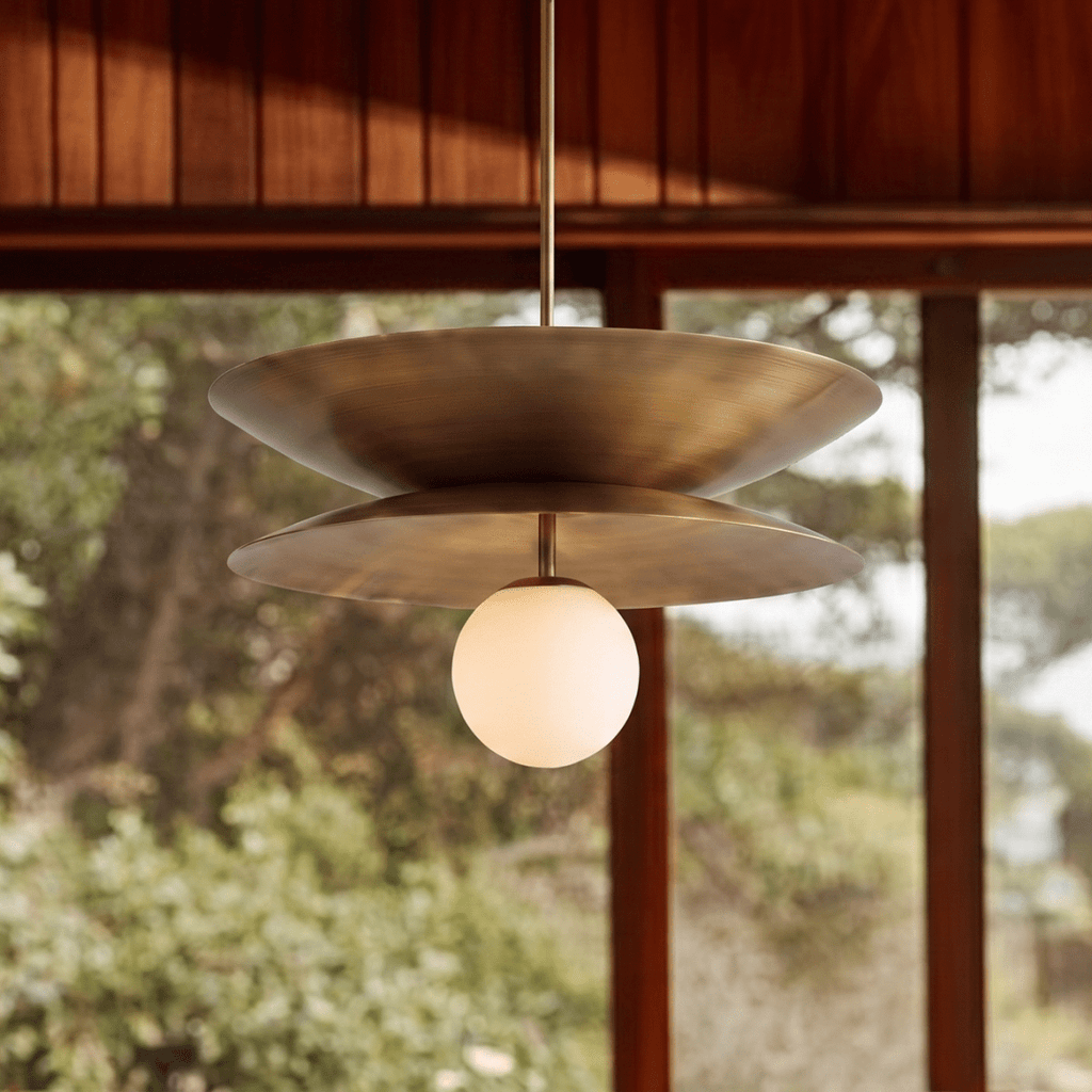 Clérachic layered-disc pendant light in vintage brass, lit against a timber-framed window full of blurred greenery.