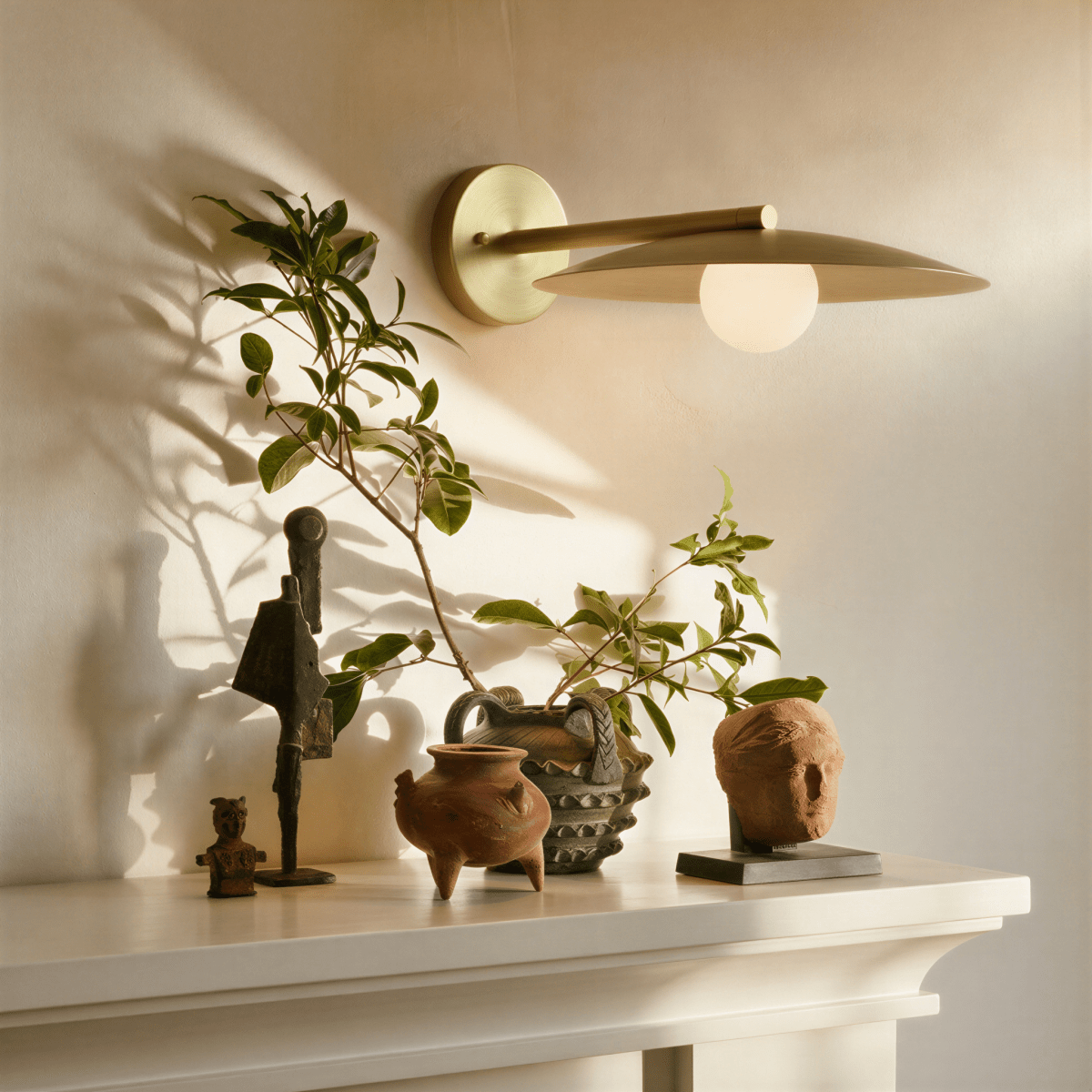 Clérachic wall lamp in brass, lit above a mantel styled with small bronze figures, terracotta vessels and a trailing plant.