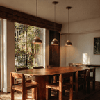 Three Clérachic pendant lights hung at staggered heights over a long oval timber dining table, beside a tall garden window.
