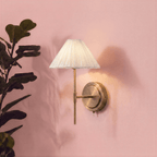 A Clérachic wall light's pleated shade glows softly on a blush pink wall beside a leafy potted fig plant.
