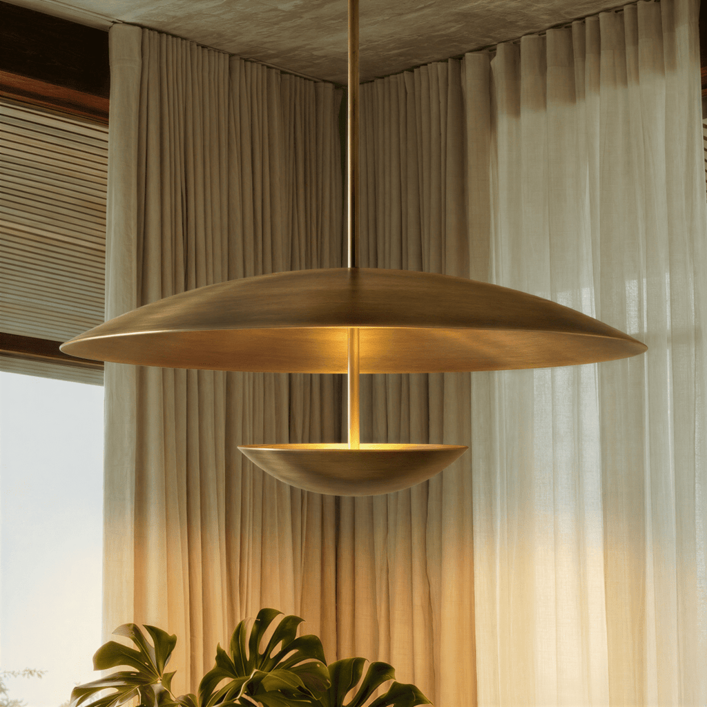 Clérachic brass pendant light in front of sheer curtains, its domed top and warm inner face both in view above a monstera plant.
