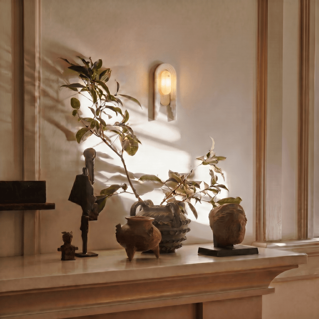 Clérachic alabaster wall light glowing in a panelled alcove above a moulded mantel shelf arranged with bronze figures, terracotta pots and an upright leafy branch.
