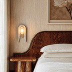 Clérachic alabaster wall light mounted bedside on a grasscloth wall, lit beside a curved burl-wood headboard and crisp white bed linen.