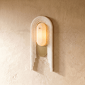 Clérachic alabaster wall light lit on a warm plaster wall, its stepped stone arch framing a glowing oval lens above a brass-finish backplate.