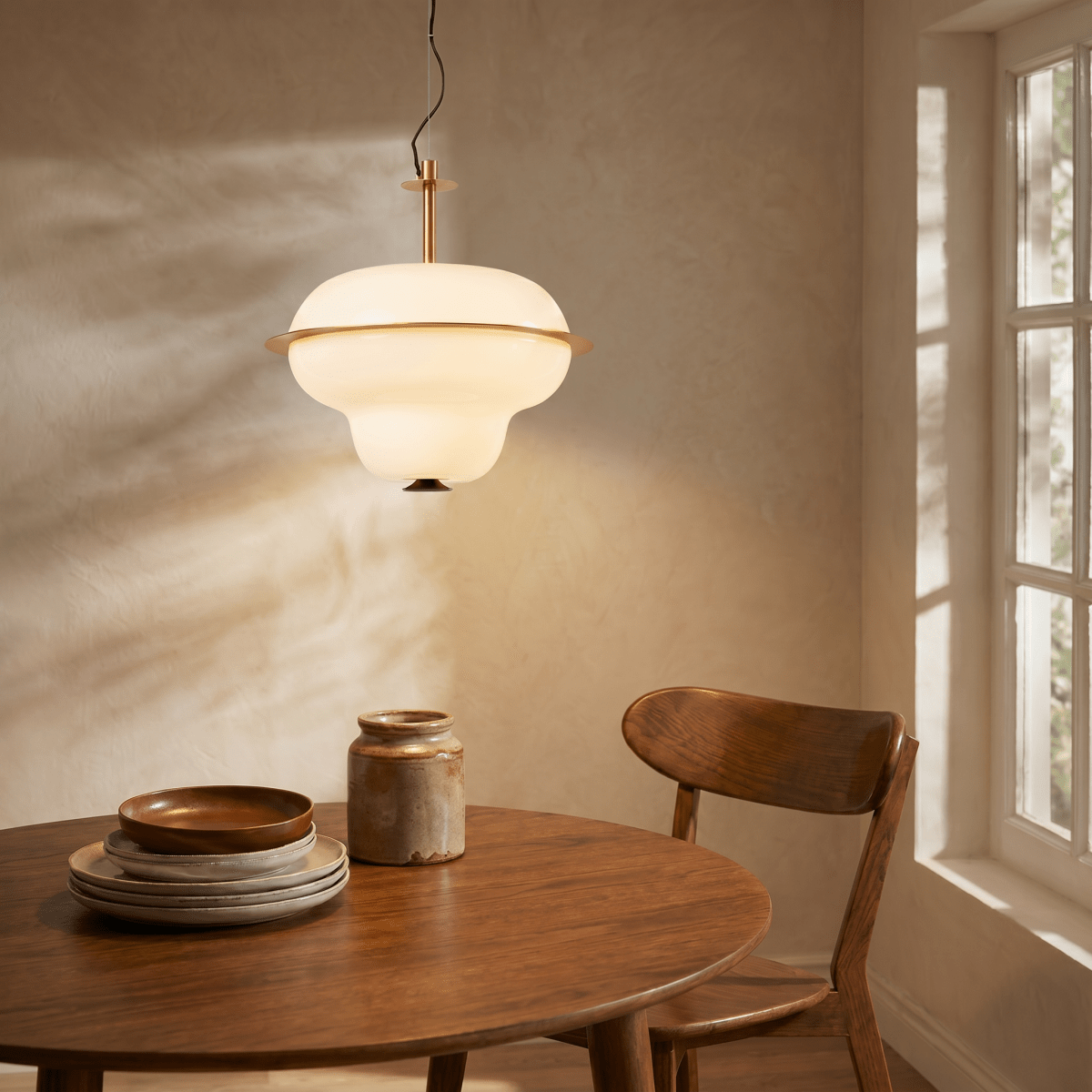 A Clérachic pendant light glows above a round wooden dining table set with rustic plates and a jug.