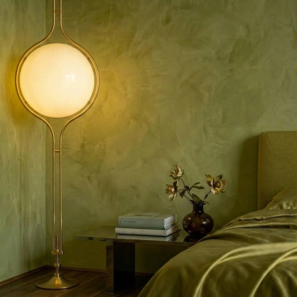 Clérachic floor-to-ceiling lamp in brass, lit against a moss-green wall beside a bedside table, the exposed threaded screw and hex nuts above its flared floor foot in view.